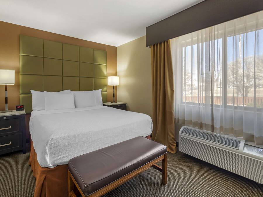 A hotel room with a large bed, bedside tables, a bench, a window with curtains, and an air conditioning unit.