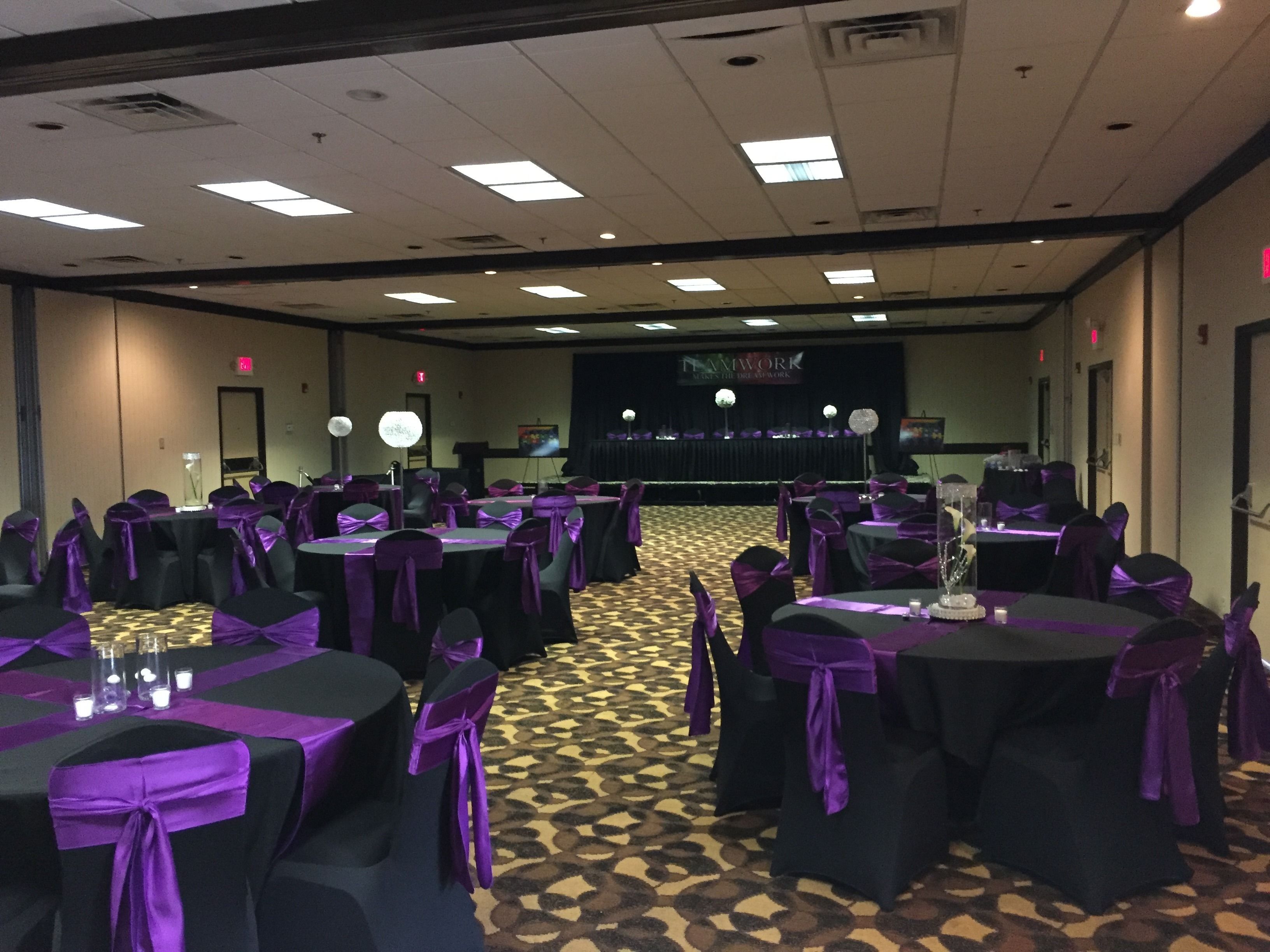 Une salle de banquet avec des tables rondes et des chaises, chacune étant ornée d’un ruban violet. La pièce est dotée d’un tapis à motifs et d’une scène sur laquelle est accrochée une banderole en arrière-plan.