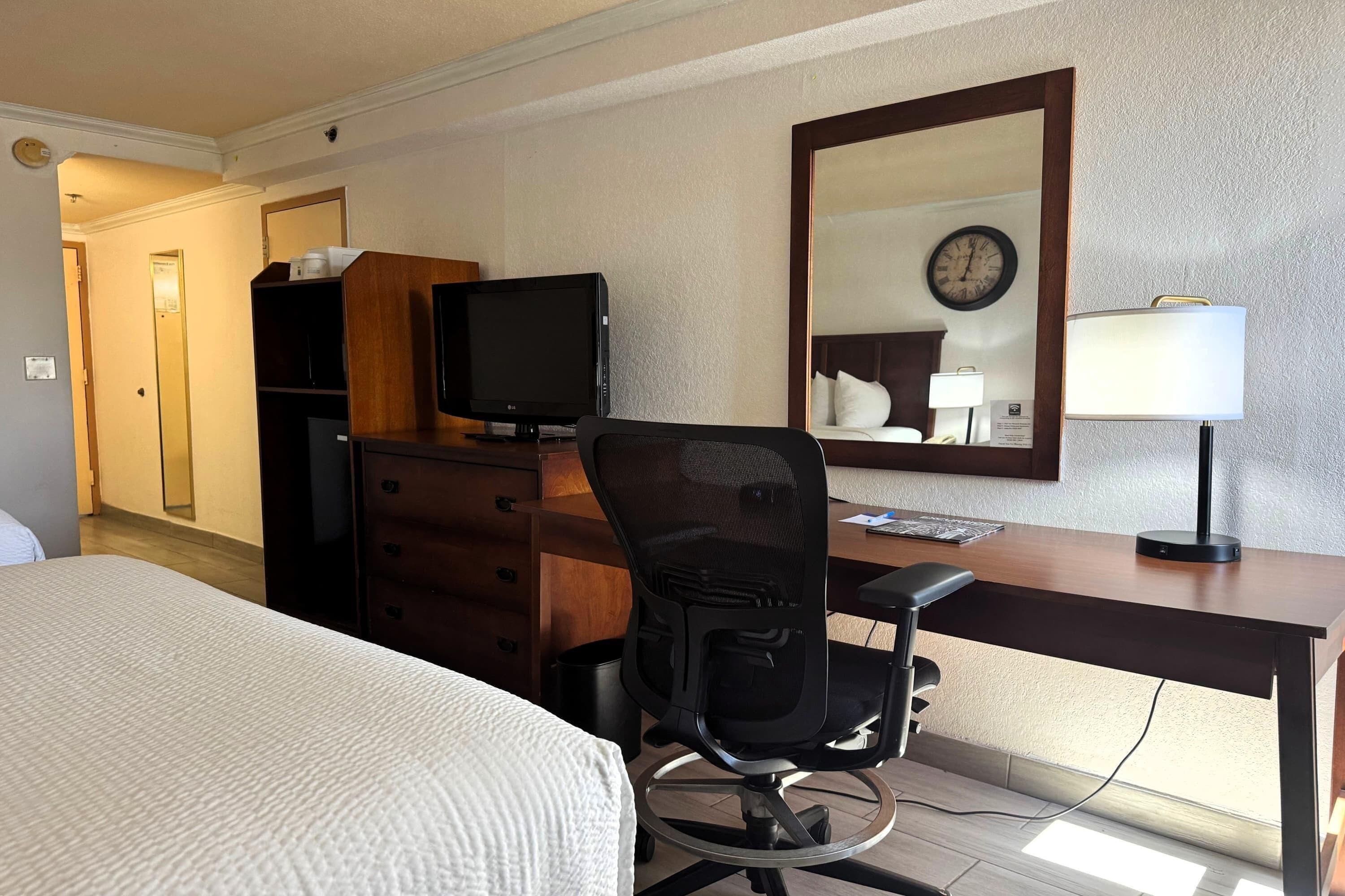 A hotel room with a bed, a desk with a computer, a chair, a television, a mirror, and a clock on the wall.