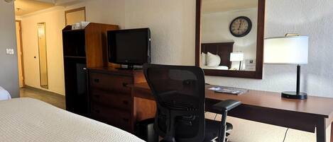 A hotel room with a bed, a desk with a computer, a chair, a television, a mirror, and a clock on the wall.