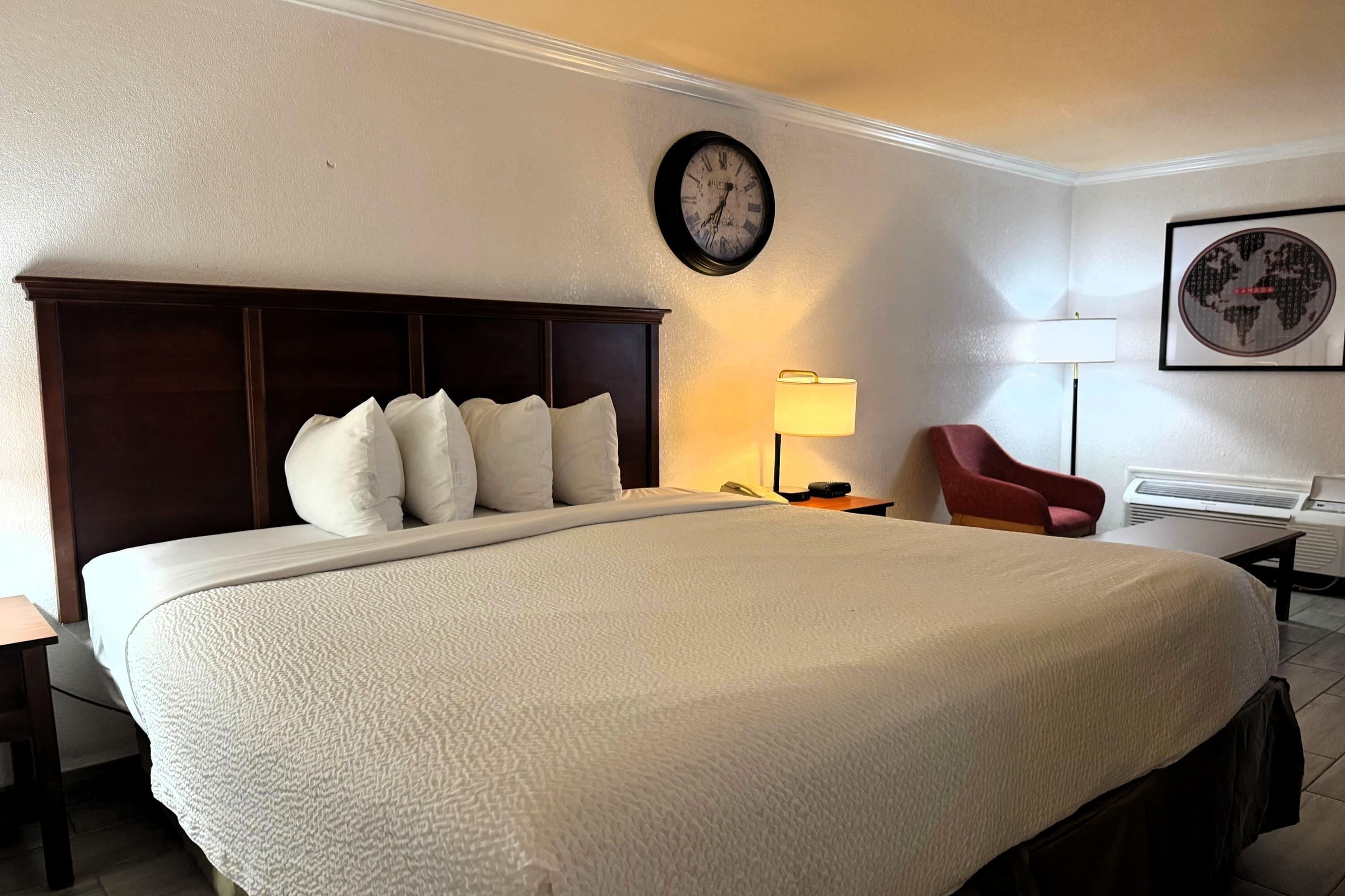 A hotel room with a large bed, a clock on the wall, a lamp, a chair, and a framed picture.
