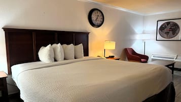 A hotel room with a large bed, a clock on the wall, a lamp, a chair, and a framed picture.