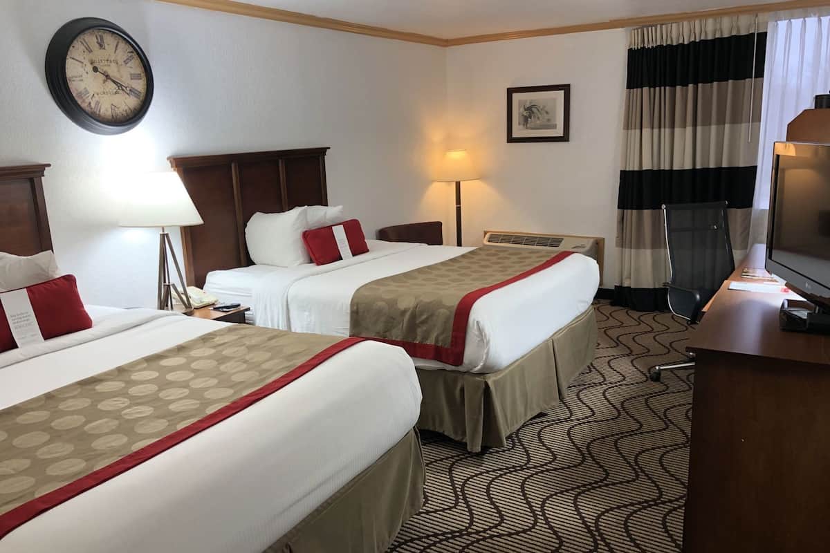Une chambre d’hôtel avec deux lits, un bureau, une télévision, une horloge et une fenêtre avec des rideaux.