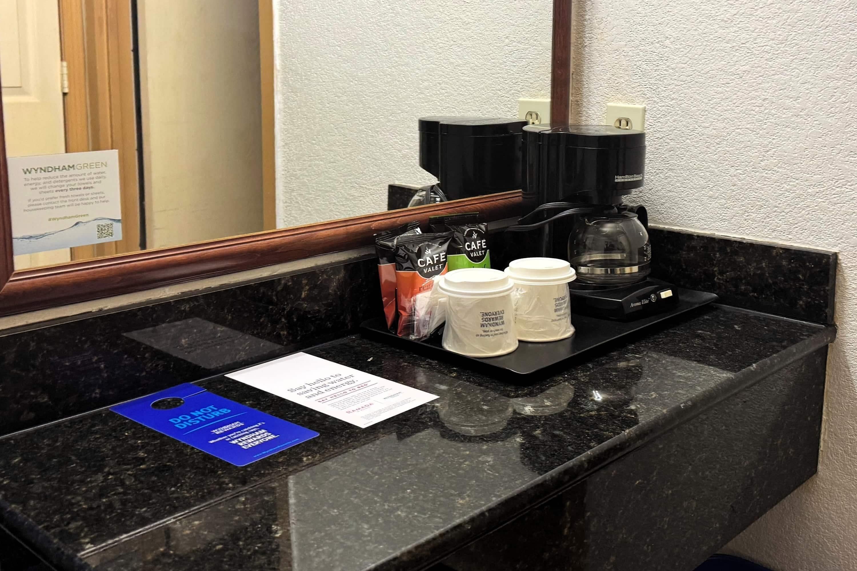 A hotel room counter with a coffee maker, disposable cups, and a sign indicating the room's name.