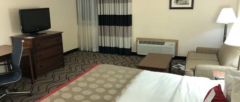 A hotel room with a bed, a desk with a computer, a chair, a TV, a lamp, and a heater.