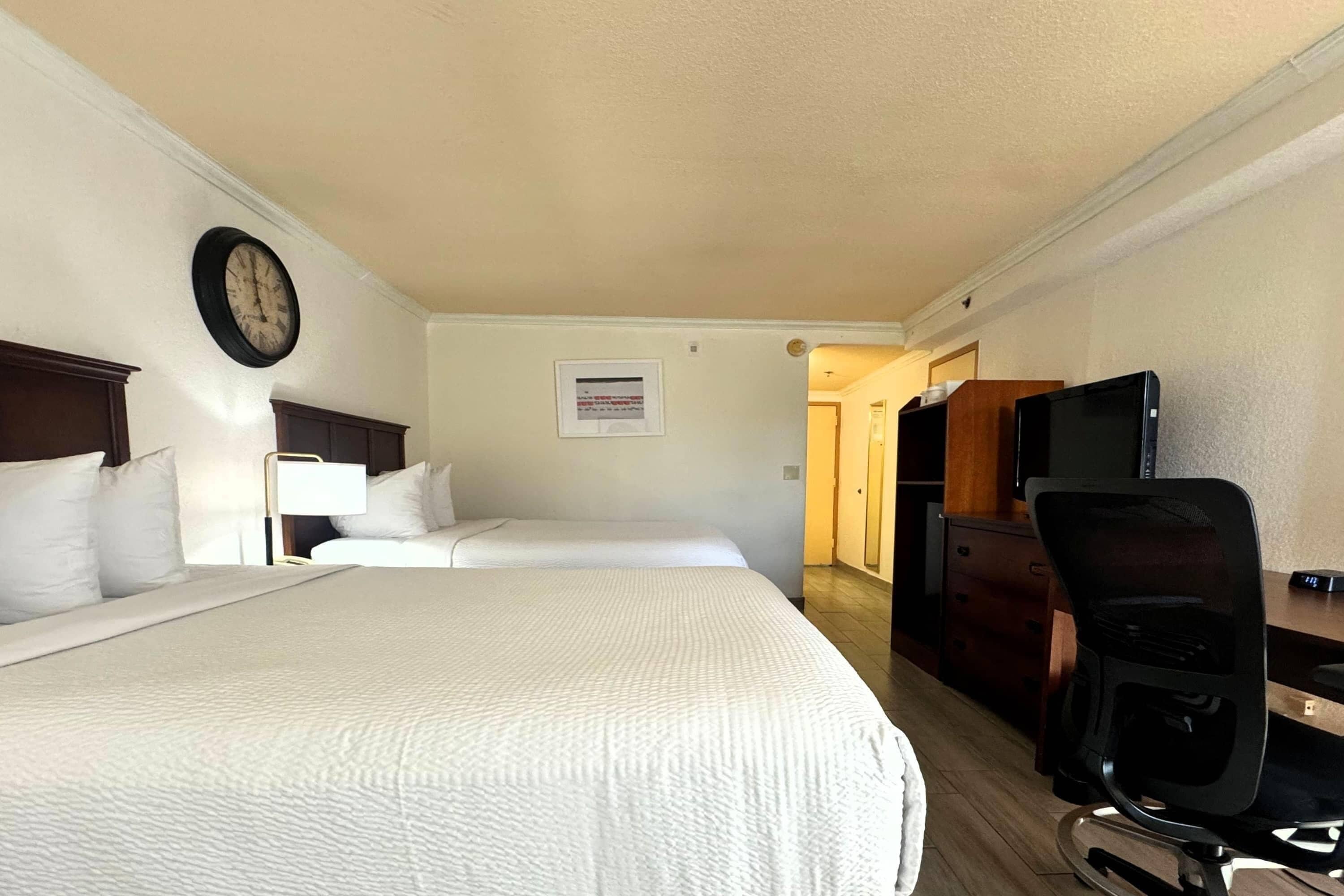 Une chambre d’hôtel avec deux lits, un bureau, une chaise, une horloge et un téléviseur.
