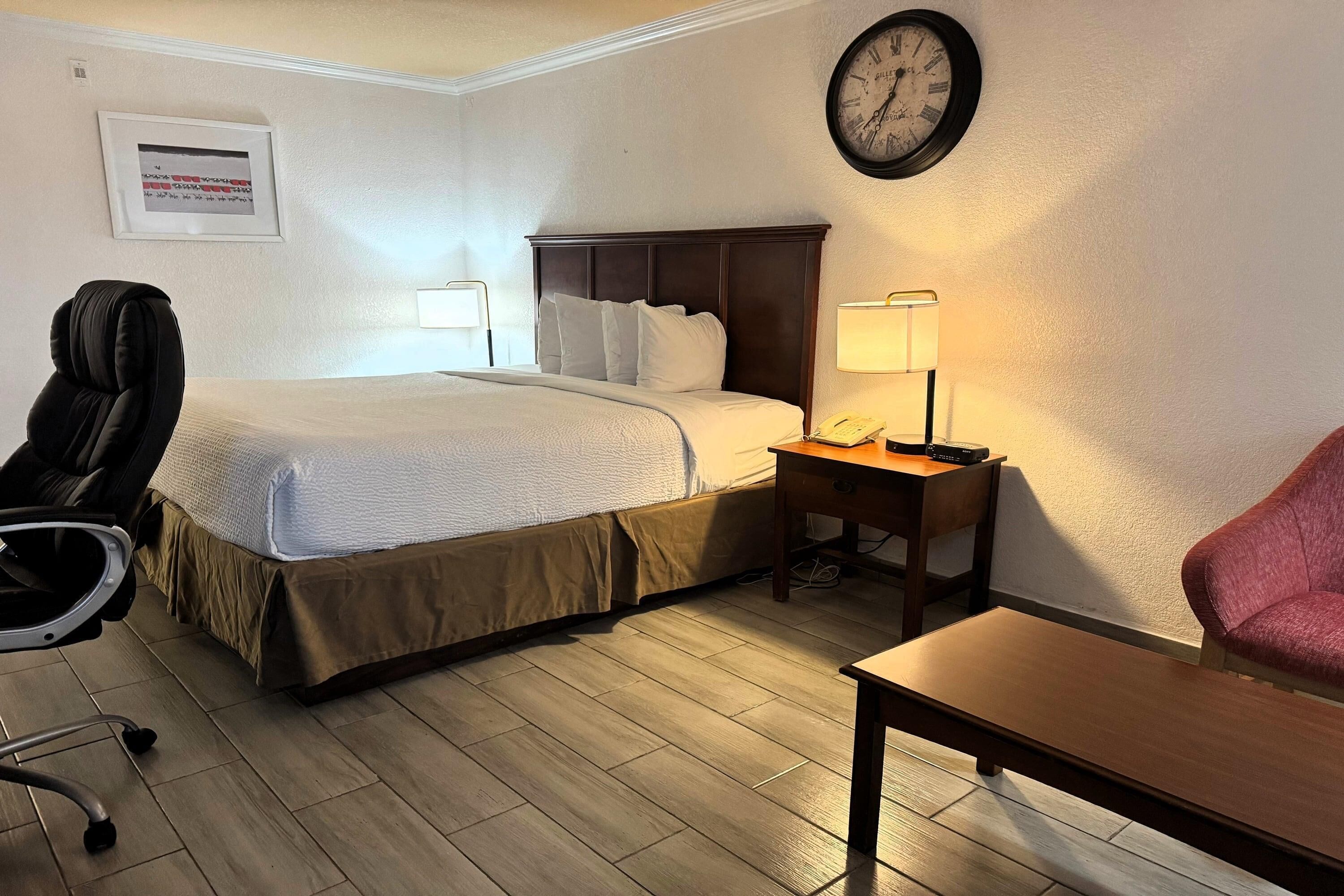 Une chambre d’hôtel avec un lit, une table de chevet avec une lampe, une chaise, un bureau et une horloge au mur.