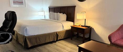 Habitación de hotel con cama, mesita de noche con lámpara, una silla, un escritorio y un reloj en la pared.