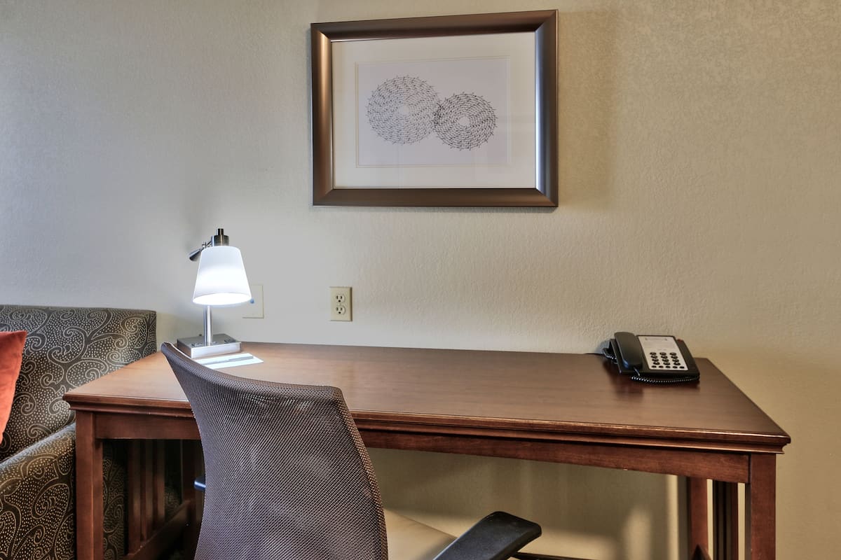 A hotel room with a wooden desk, a chair, a lamp, a framed artwork, and a telephone.