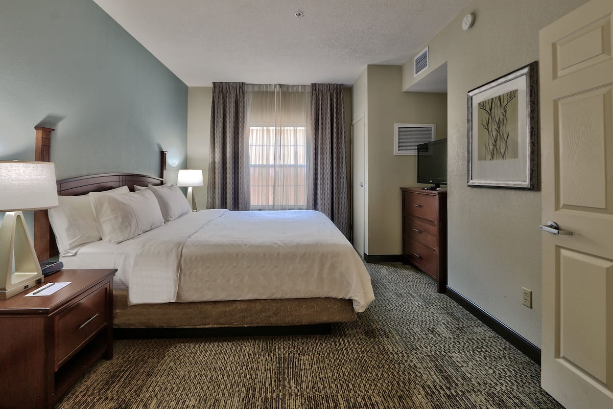 A hotel room with a bed, a nightstand, a lamp, a dresser, a framed picture on the wall, and a window with curtains.