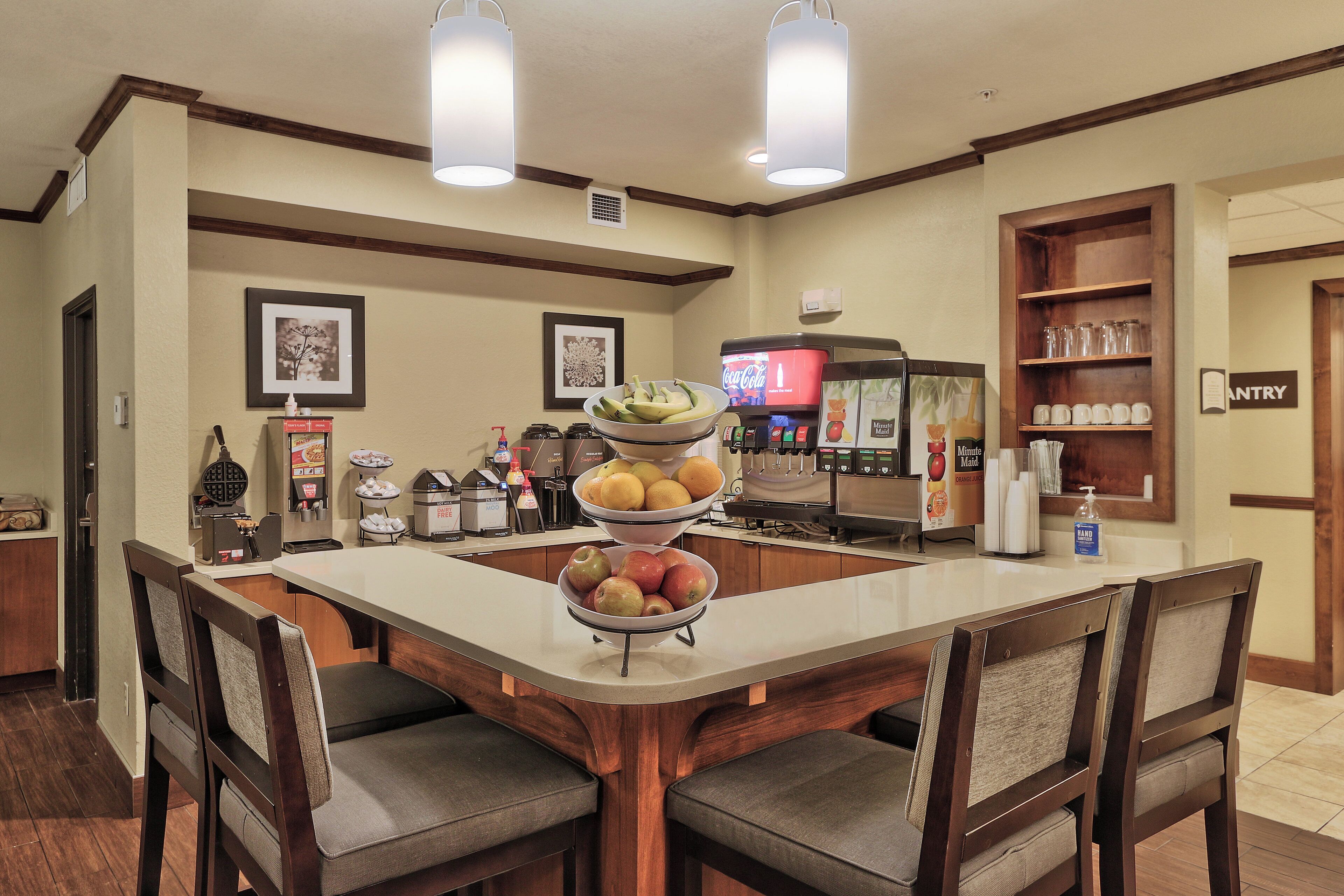 A hotel dining area with a central island, chairs, a beverage dispenser, and a refrigerator.