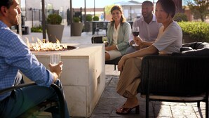 A group of people sitting around an outdoor fire pit with a table, in a patio area with chairs and potted plants.