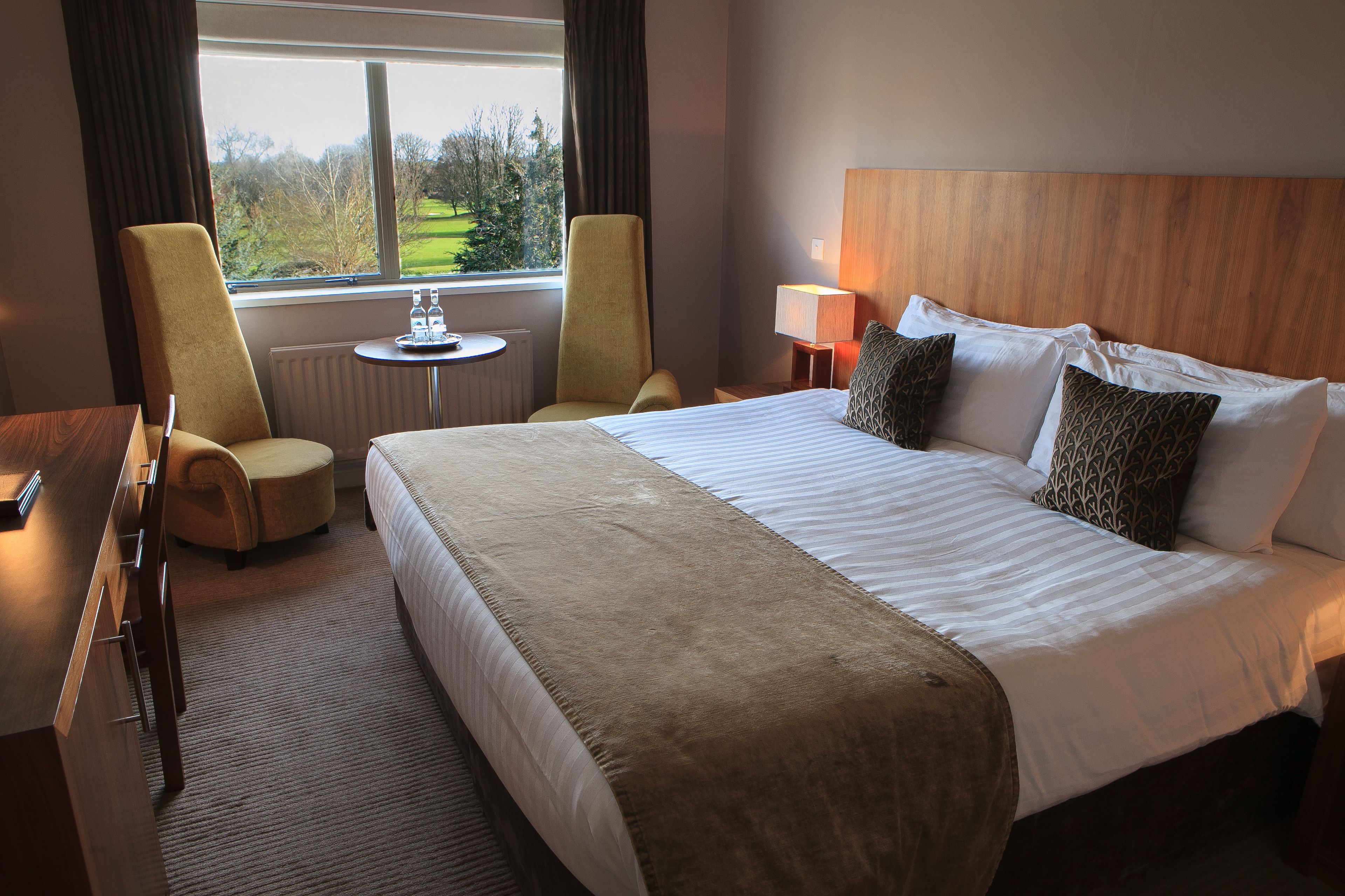 Habitación de hotel con una cama grande, dos sillones, una mesita de noche con lámpara y vistas al exterior a través de una ventana.