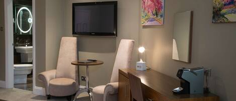 A hotel room with a TV, two armchairs, a desk with a coffee machine, and artwork on the walls.