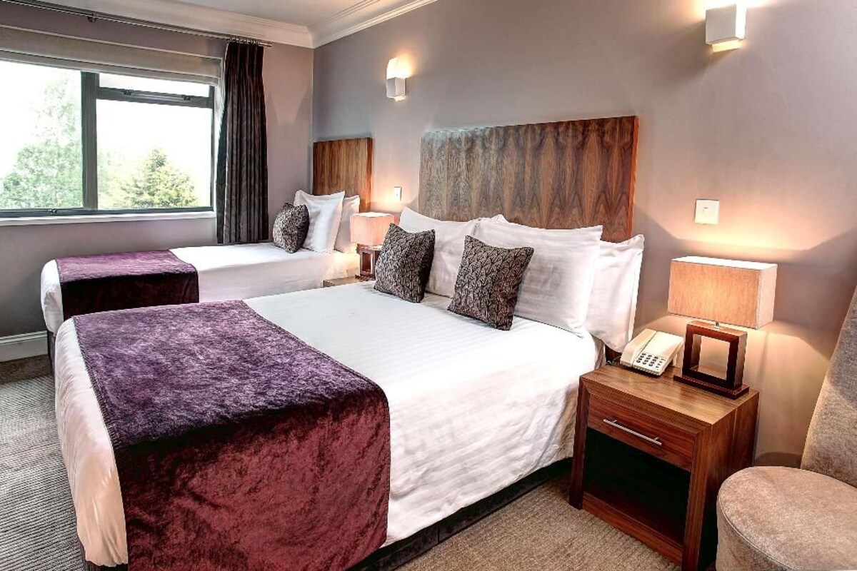 A hotel room with two beds, a nightstand with a lamp, and a view of the outdoors.