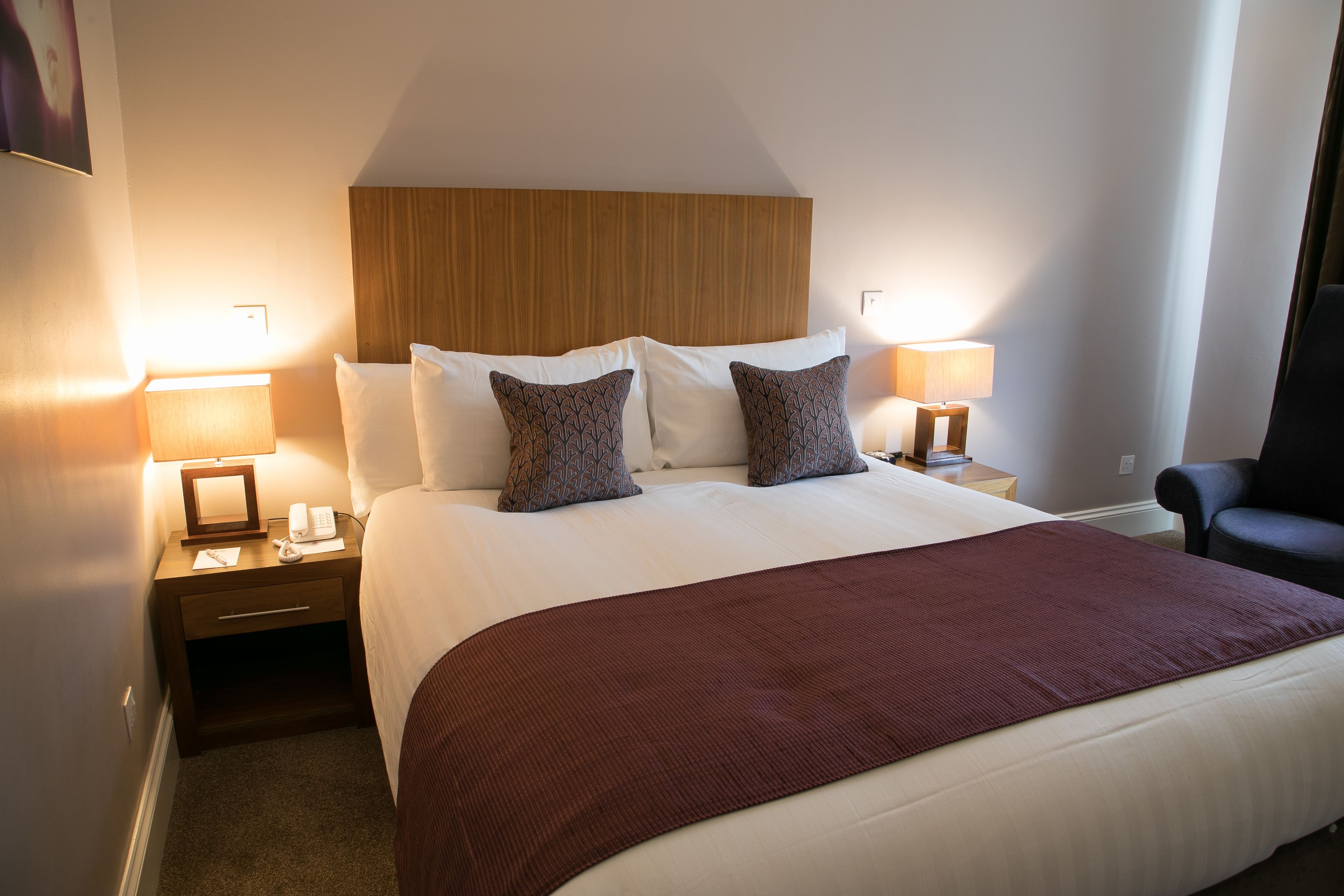 A hotel room with a large bed, two bedside tables with lamps, a wooden headboard, and a dark armchair.