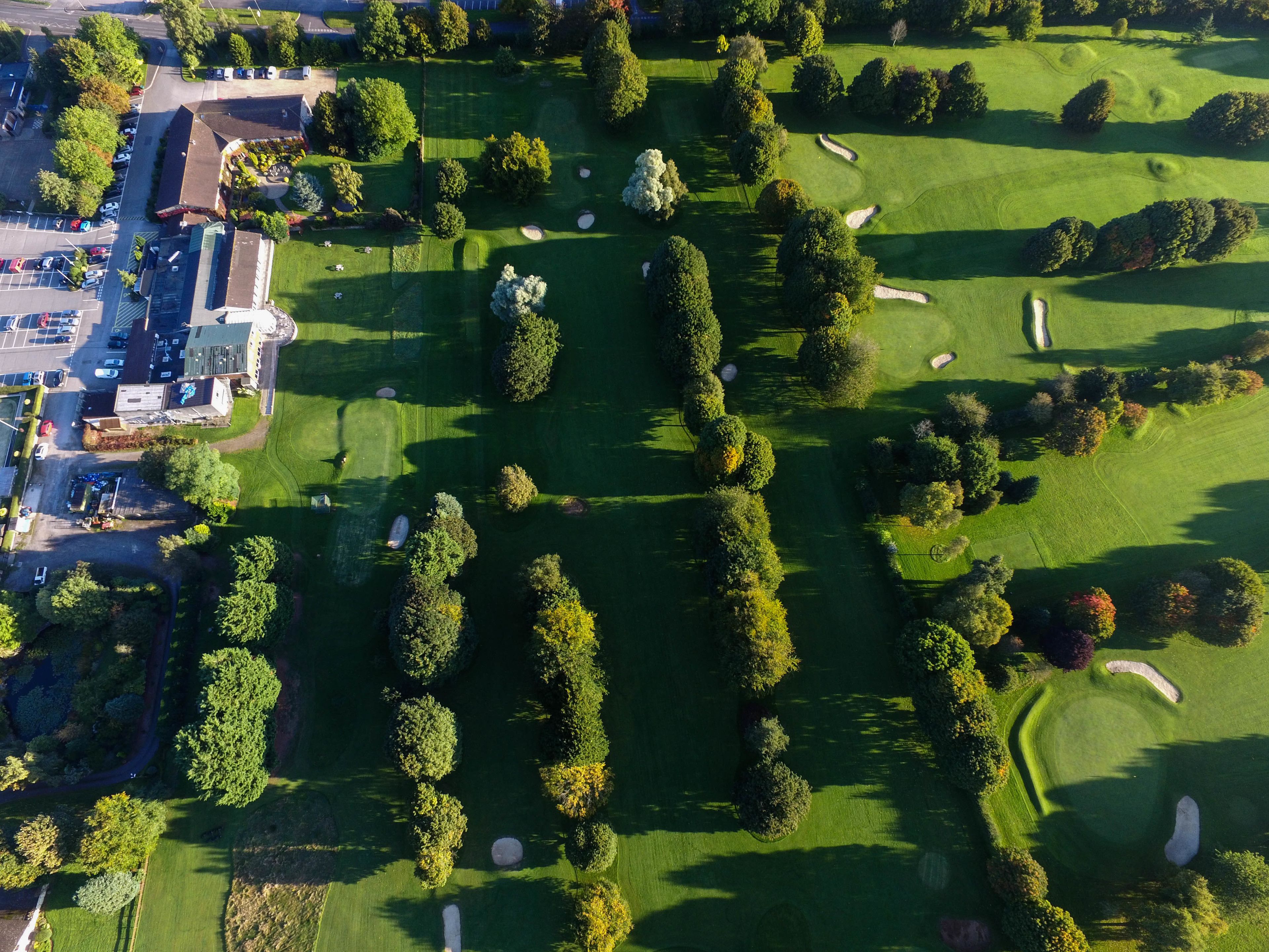 An aerial view of a golf course with neatly trimmed trees, sand bunkers, and a parking area with buildings.