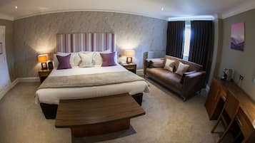 A hotel room with a large bed, a sofa, a wooden coffee table, and a desk with a coffee machine.