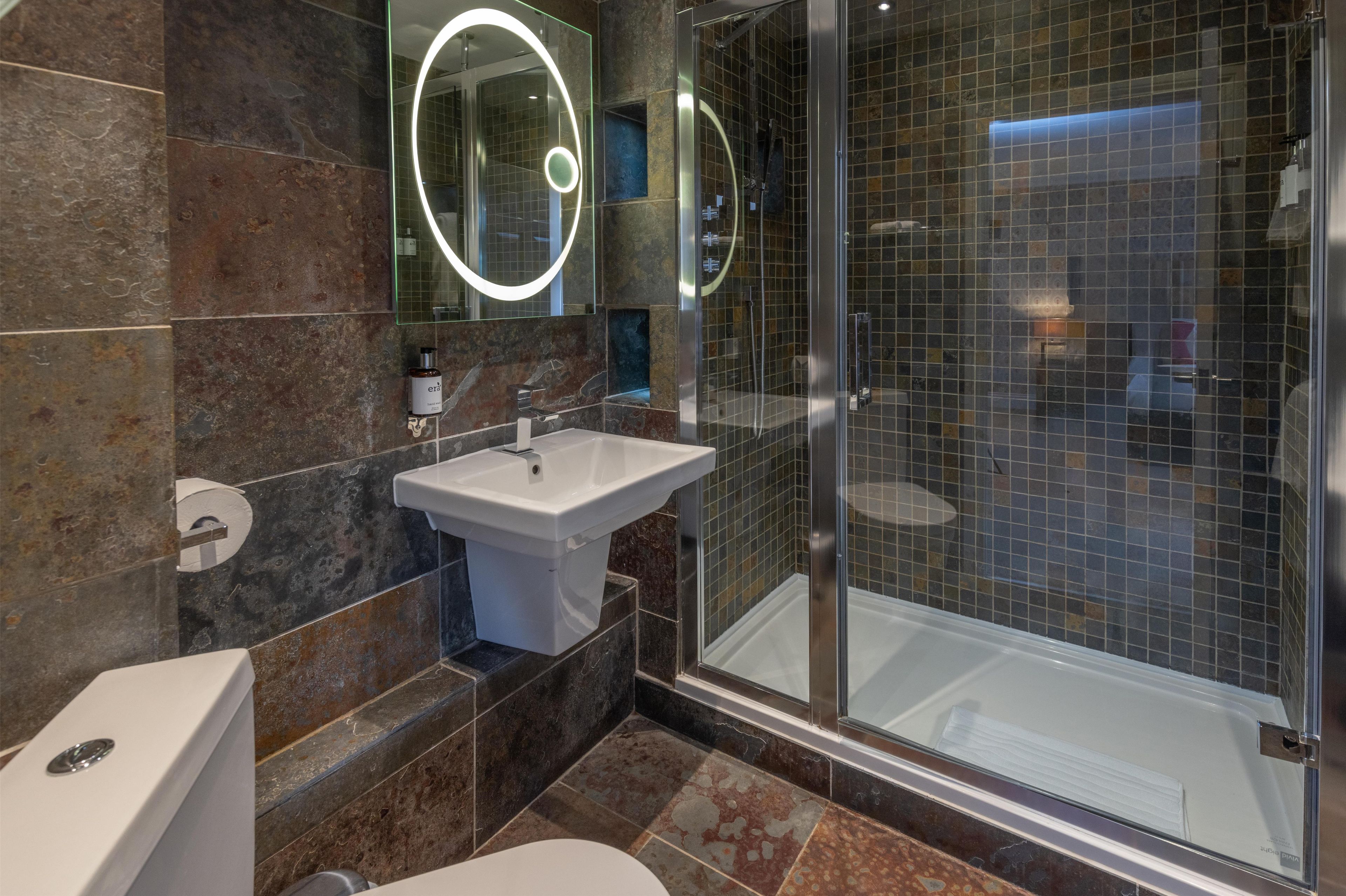 A modern bathroom with a glass-enclosed shower, a pedestal sink, and a large mirror with a circular light.