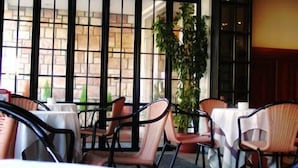 Un espacio interior con mesas y sillas, un ventanal con vista a un edificio de ladrillo, y una planta en maceta.