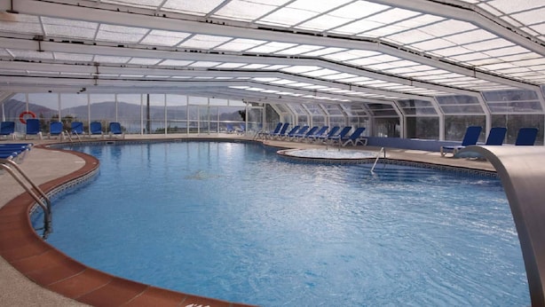 Una piscina interior con agua azul, rodeada de sillas reclinables azules y un techo de cristal.