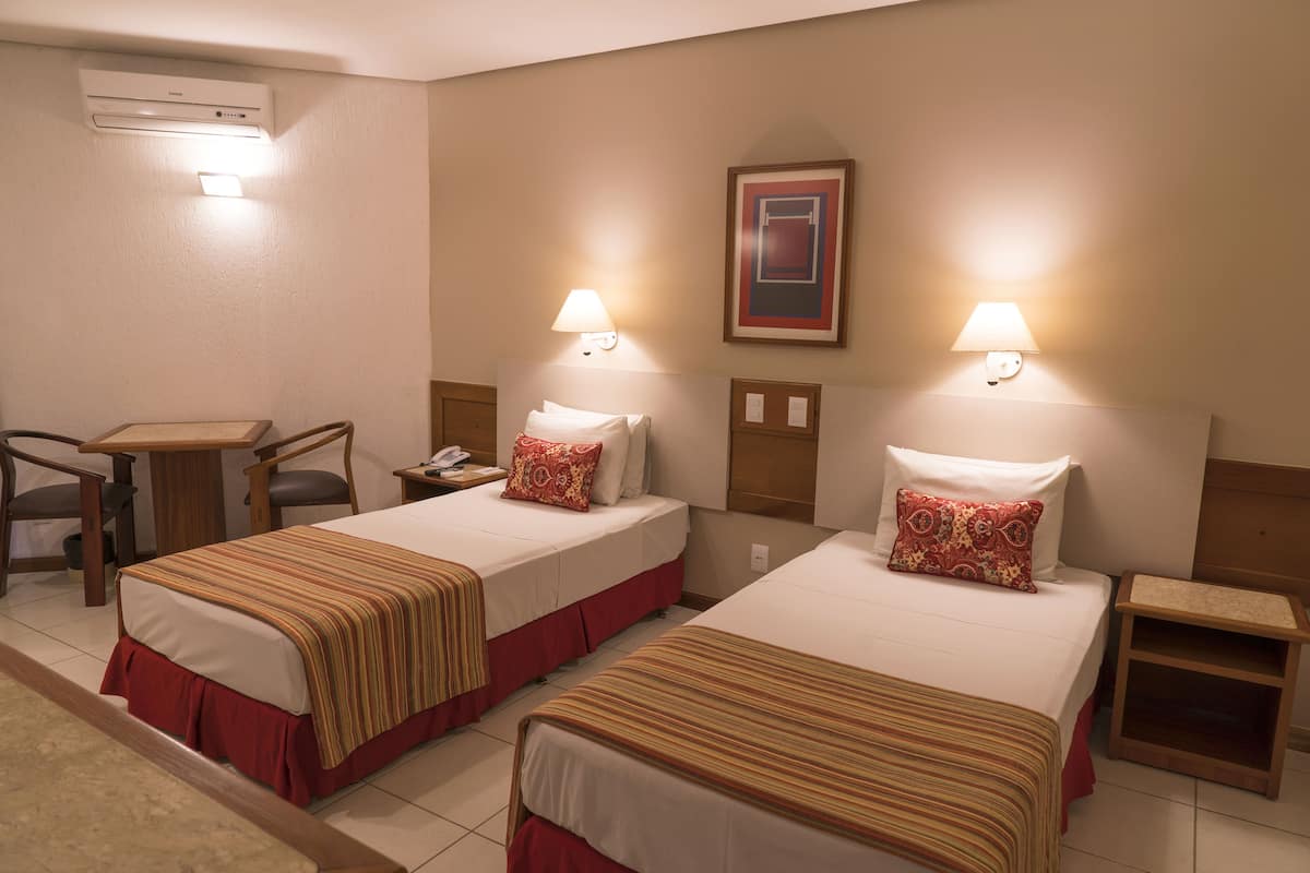 Habitación de hotel con dos camas, cada una con colcha a rayas y falda roja. Hay mesitas de noche con lámparas, un escritorio con silla y aire acondicionado de pared.