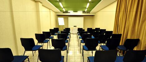 Un salón de conferencias con filas de sillas negras con cojines azules, una pantalla de proyector al frente y un techo verde.