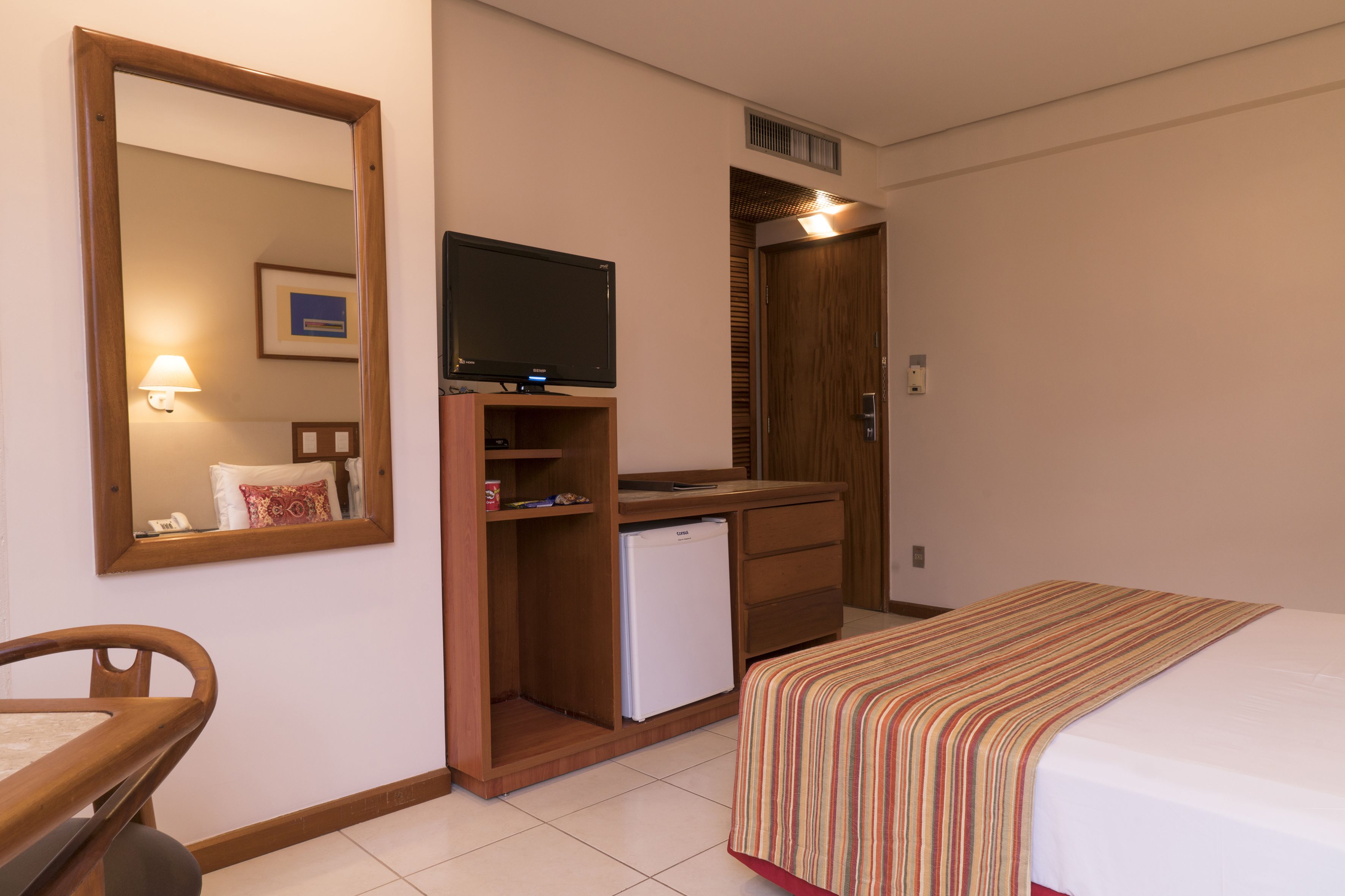 A hotel room with a bed, a desk with a computer, a small refrigerator, a mirror, and a television.