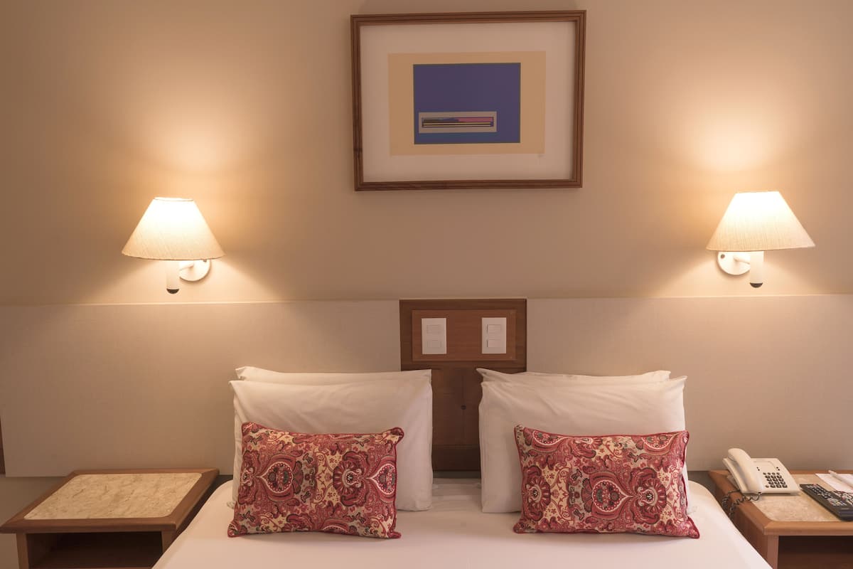 Habitación de hotel con dos camas, cada una con un cojín estampado y una mesita de noche con lámpara. Encima de las camas hay un cuadro enmarcado en la pared.
