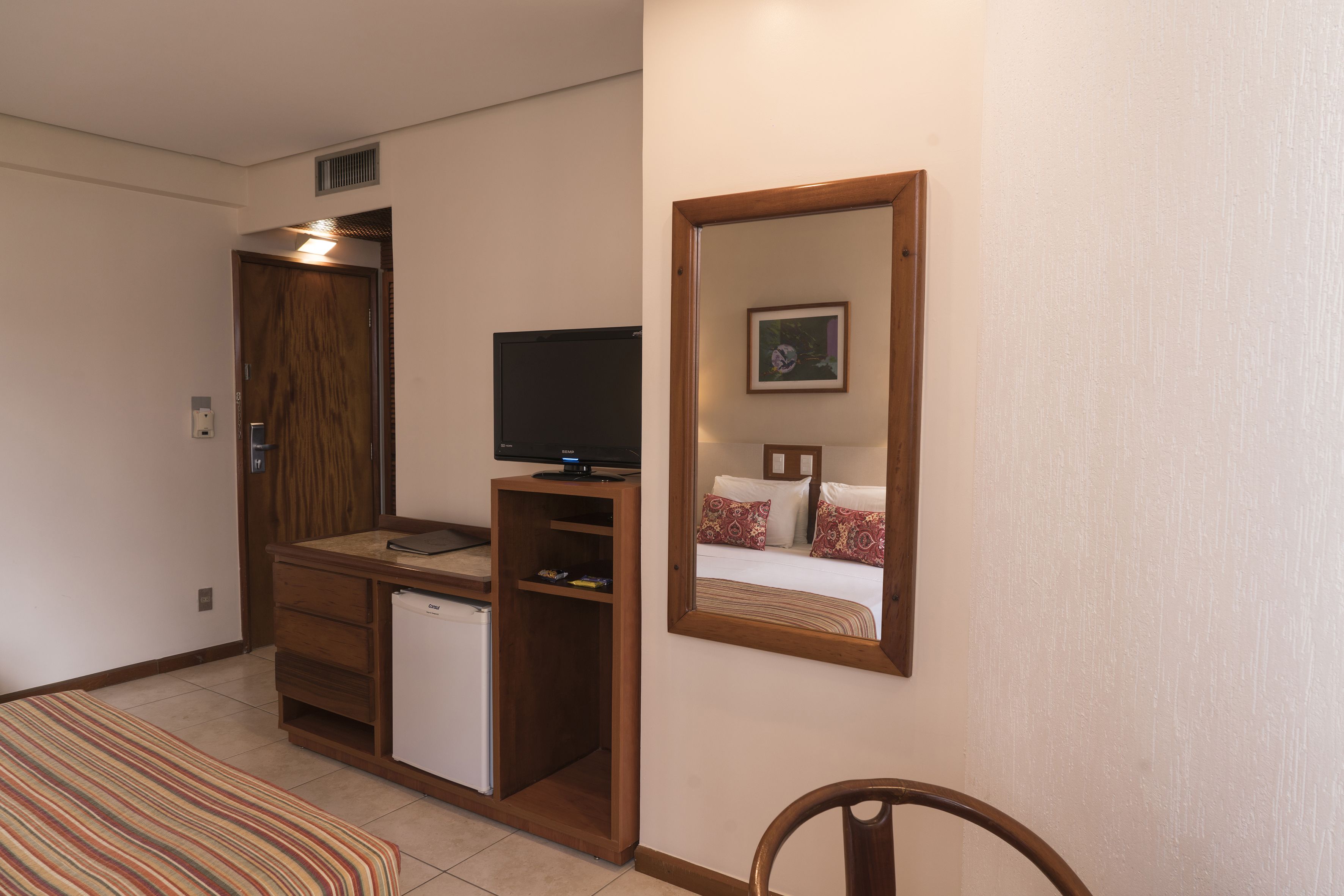 A compact hotel room with a bed, a small desk with a television, a mini-fridge, and a mirror on the wall.