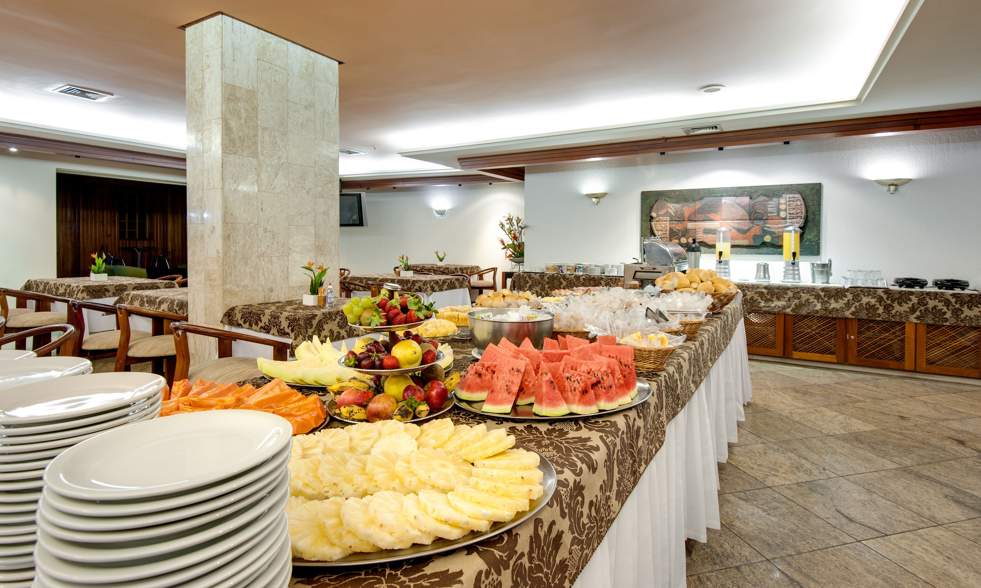 Buffet com diversas frutas, doces e um espaço de bebidas em um salão bem iluminado com colunas de mármore e teto alto.