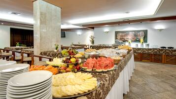 A buffet with various fruits, pastries, and a beverage station in a well-lit room with marble columns and a high ceiling.