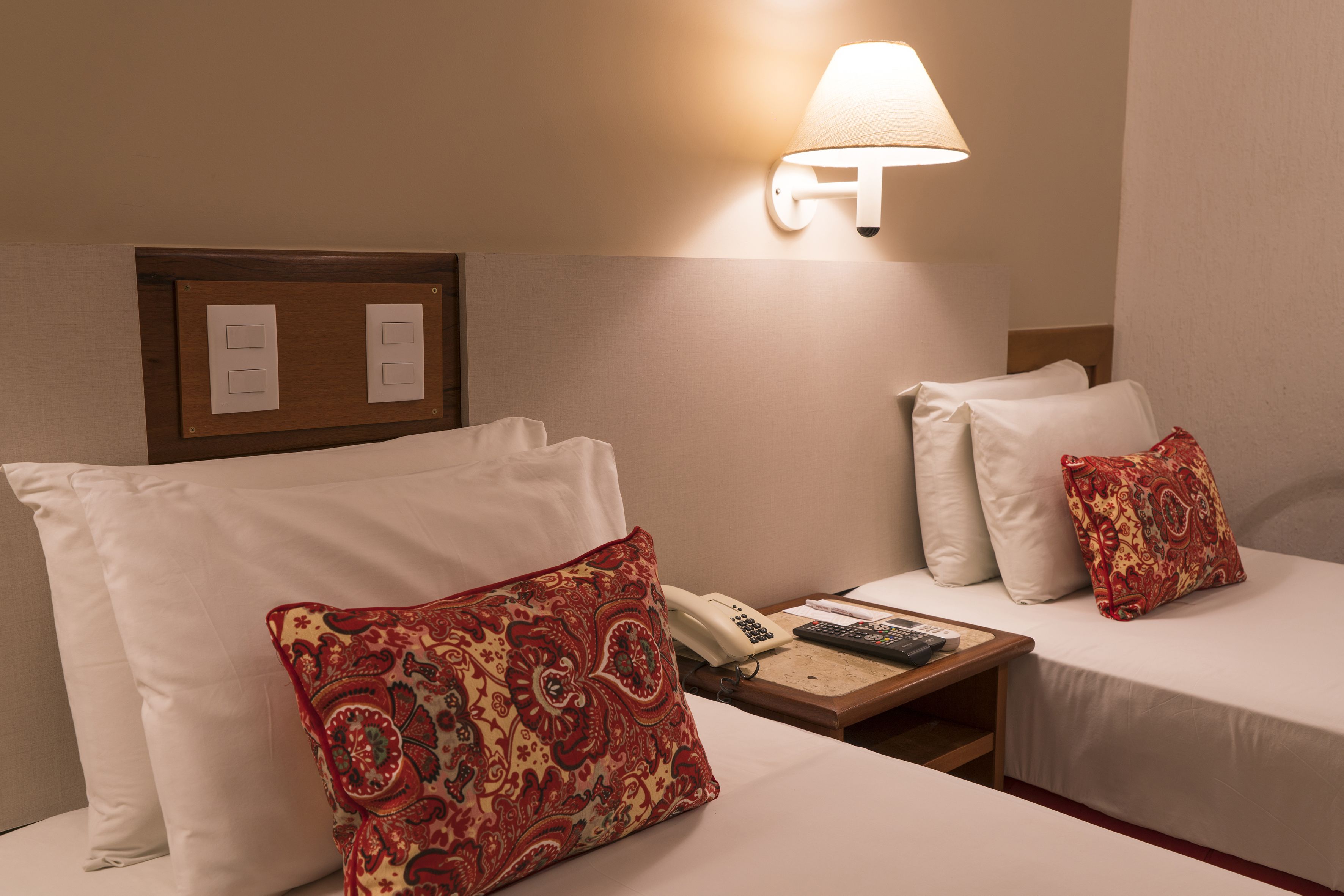 A hotel room with two beds, a bedside table with a lamp, a phone, and a remote control.