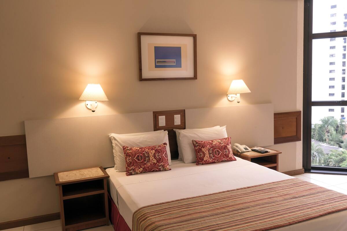 Habitación de hotel con una cama grande, dos mesitas de noche, una lámpara de noche, un cuadro en la pared y una ventana con vistas a edificios y árboles.