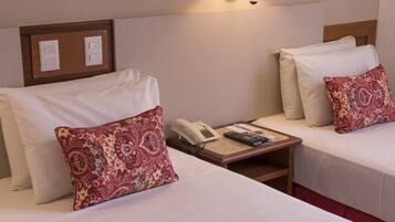 Quarto de hotel com duas camas, cada uma com lençóis brancos e almofada vermelha estampada. Tem uma mesa de cabeceira com um telefone e um controle remoto.
