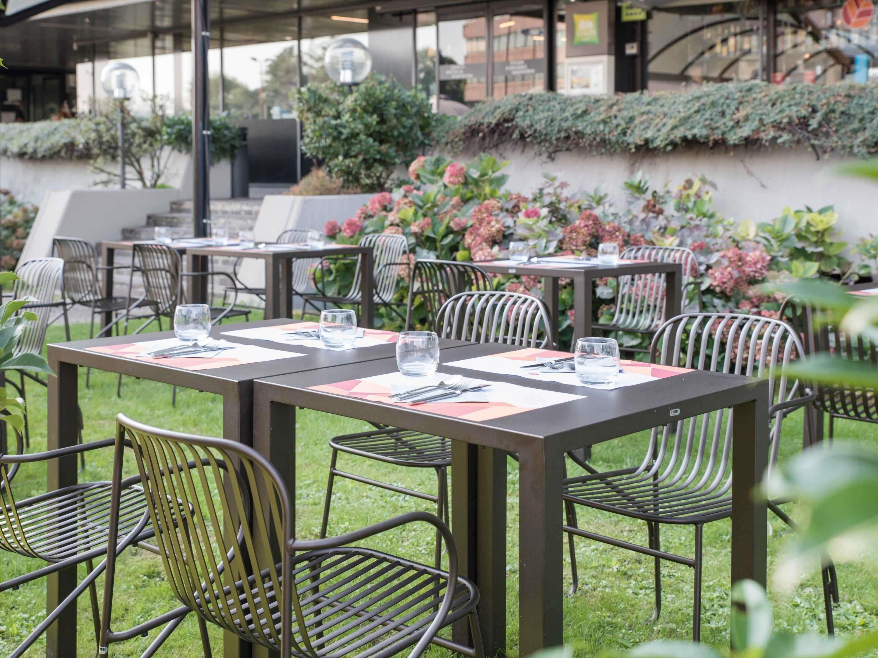 Un espacio de comedor al aire libre con mesas y sillas, rodeado de follaje y flores.