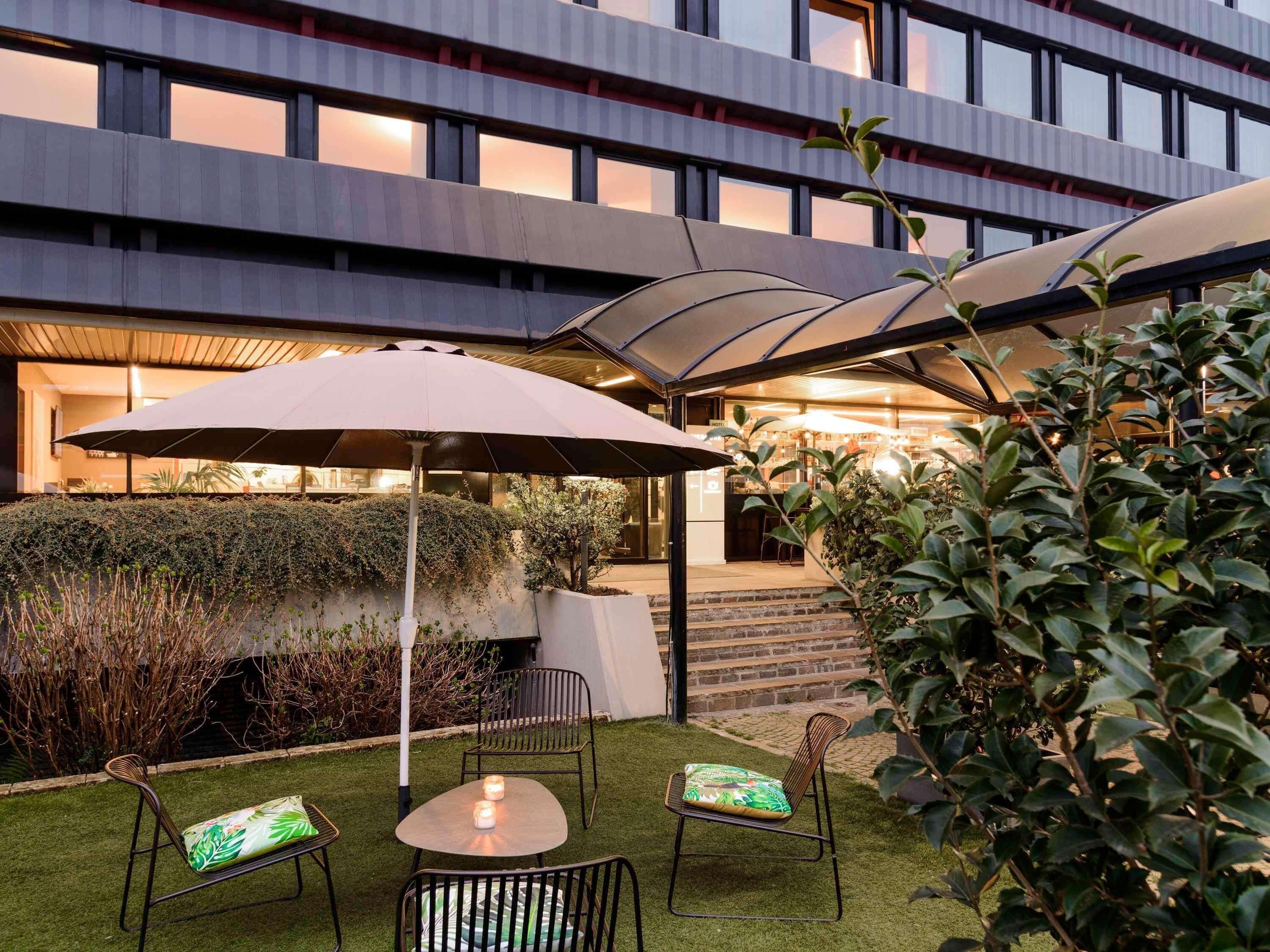 An outdoor seating area with chairs and an umbrella, surrounded by greenery and a building facade.