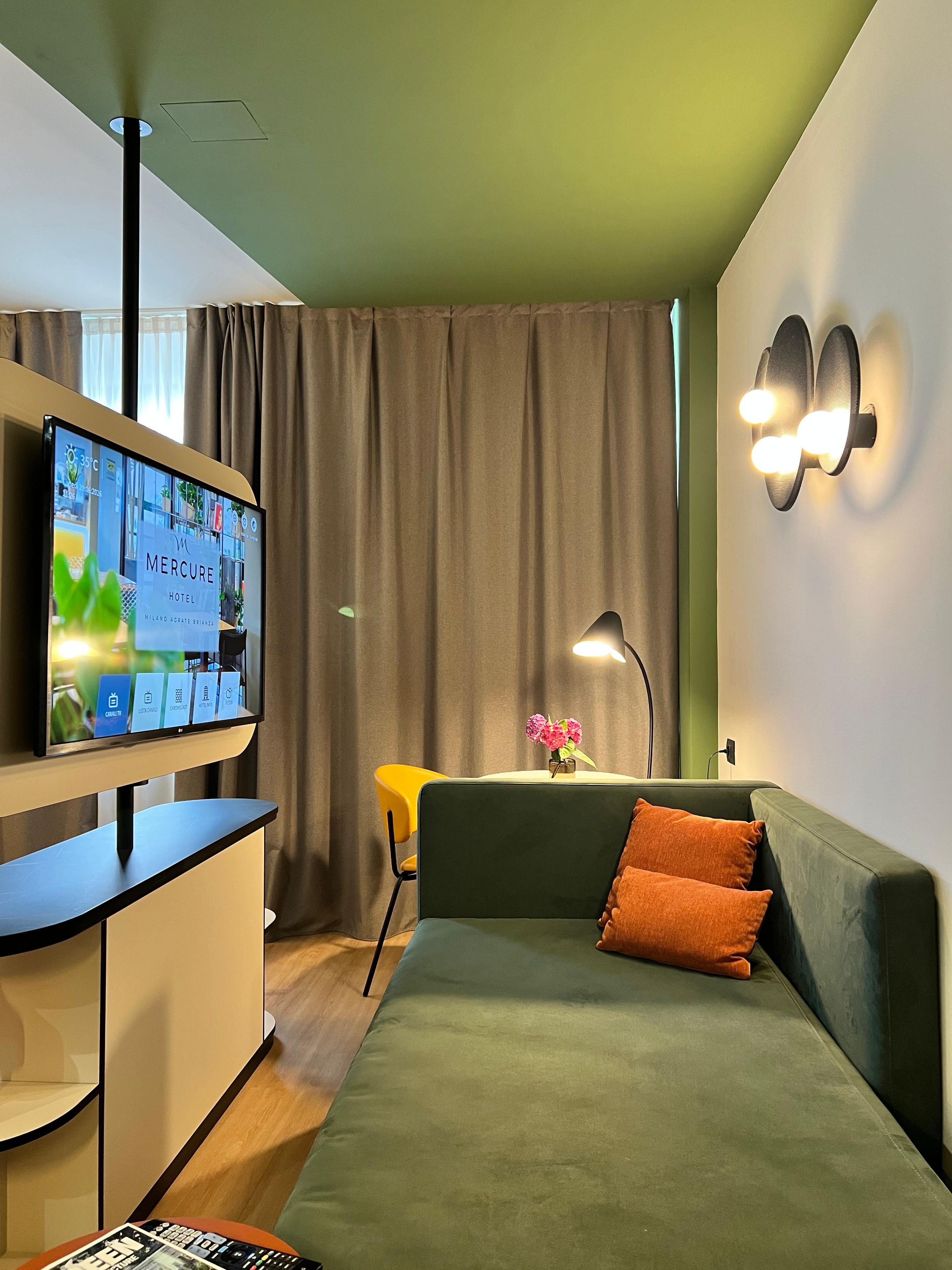 A modern hotel room with a flat-screen TV, a sofa, a small table with a vase of flowers, and a wall-mounted light fixture.