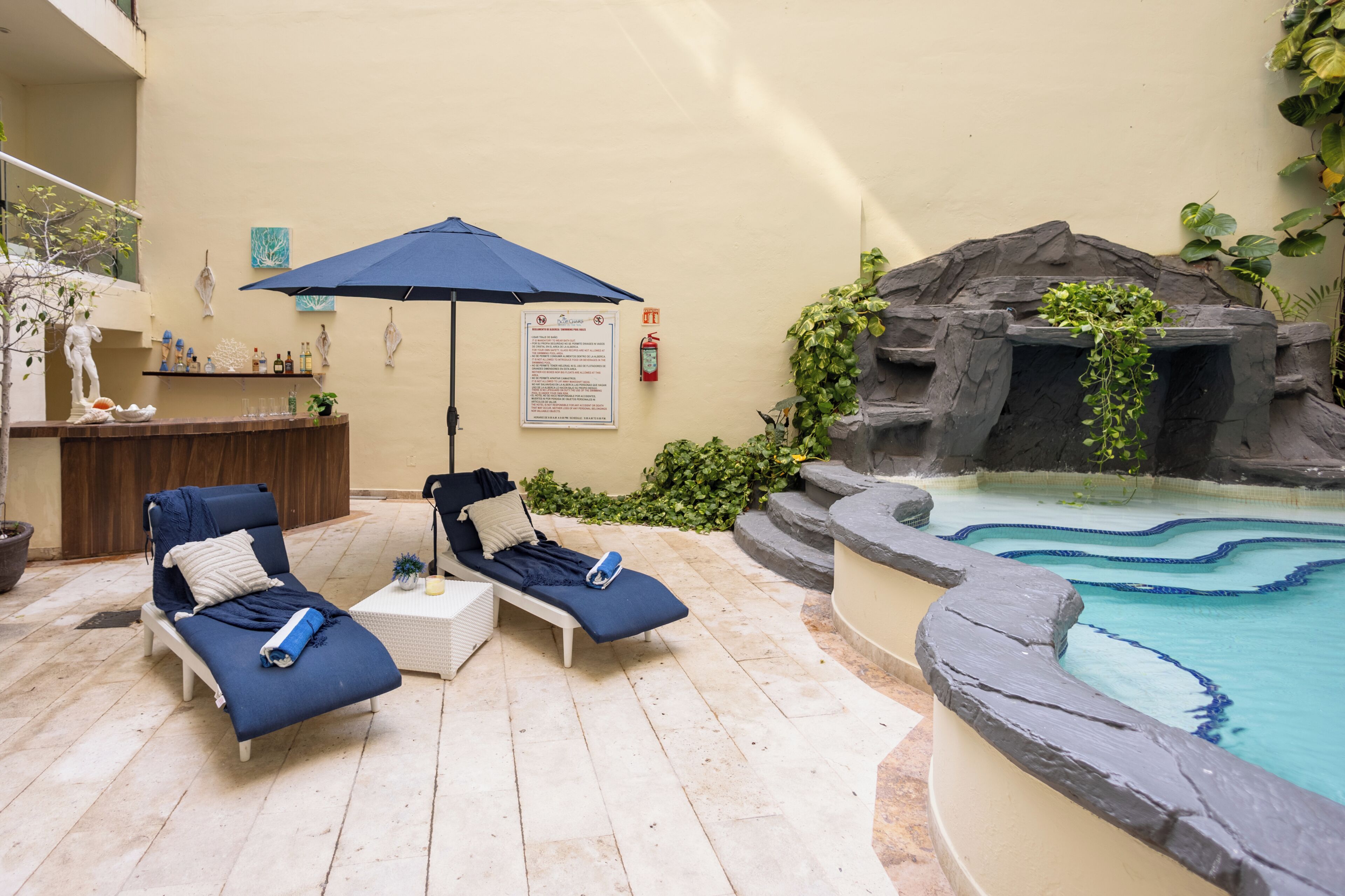 A poolside area with lounge chairs, a blue umbrella, a small bar, and a decorative rock wall.