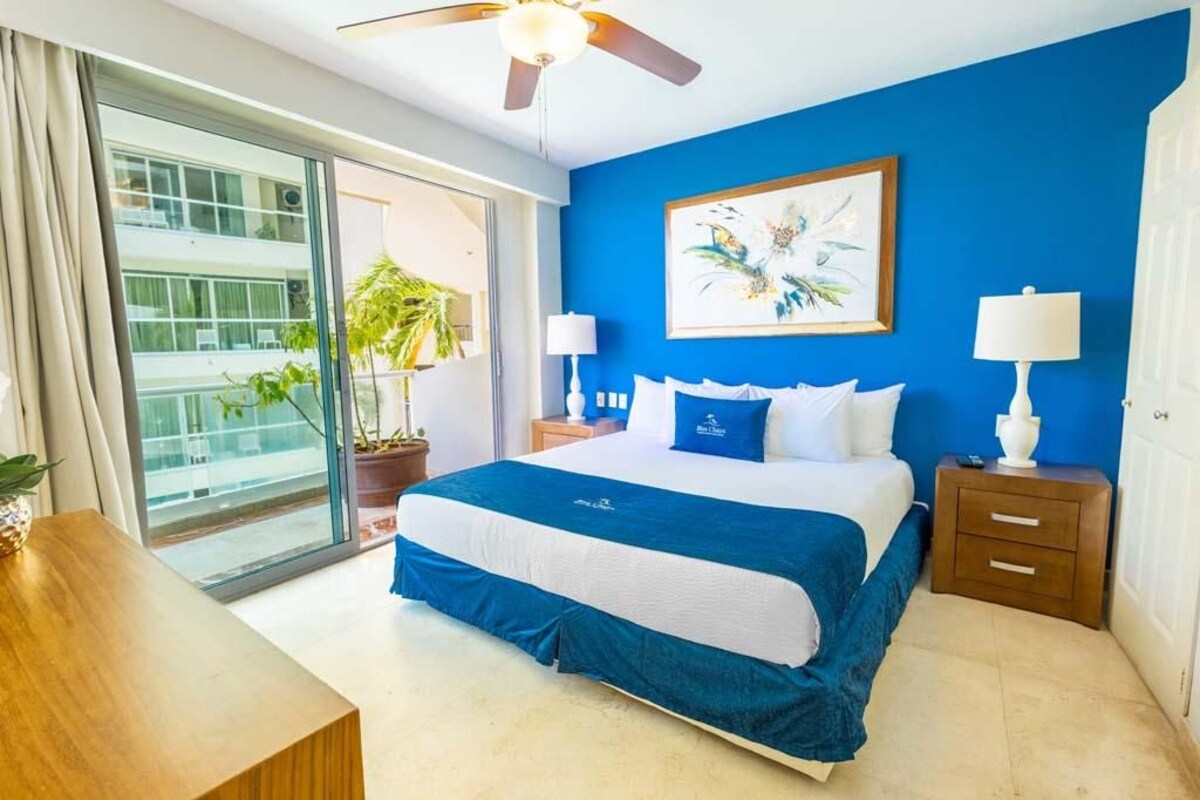 A hotel room with a large bed, a ceiling fan, a painting, and a view of a building through a glass door.