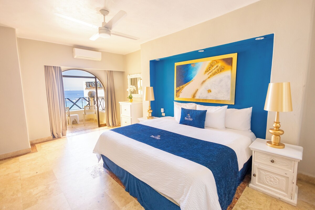 A hotel room with a large bed, a blue accent wall, a painting, two bedside lamps, and a view through a glass door.
