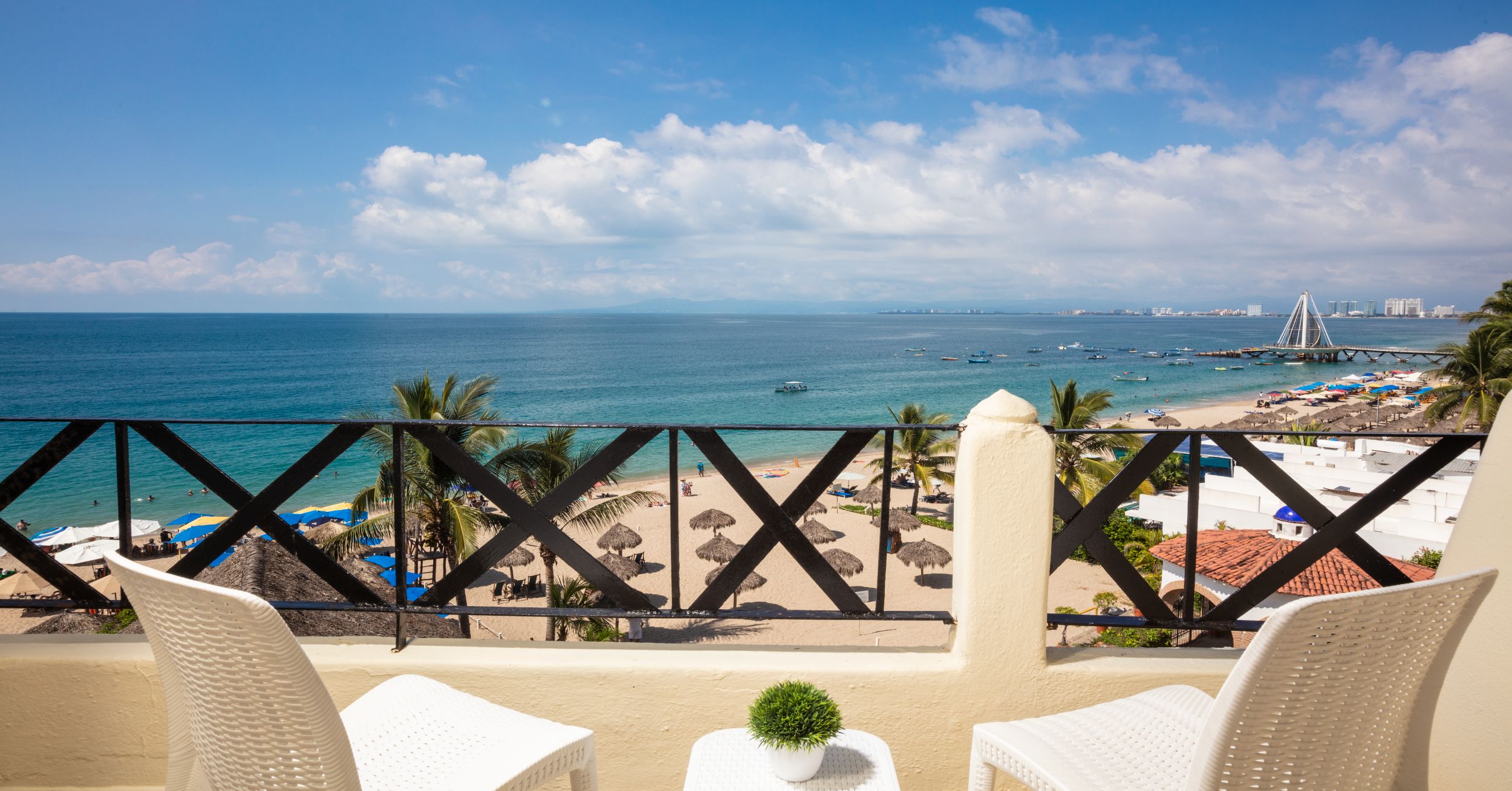 A balcony with a view of the beach, palm trees, and a distant city skyline.