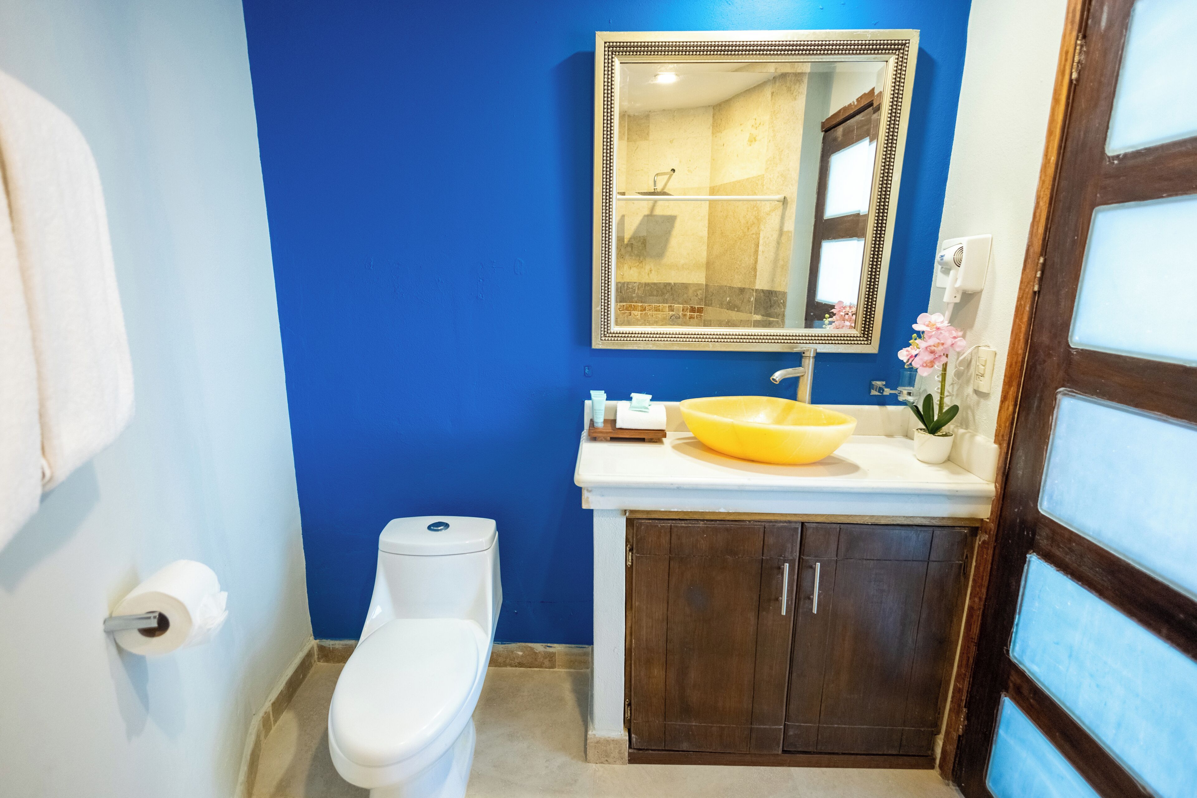 Een badkamer met een blauwe wand, een wit toilet, een geel wastafelmeubel en een spiegel boven het wastafelmeubel.