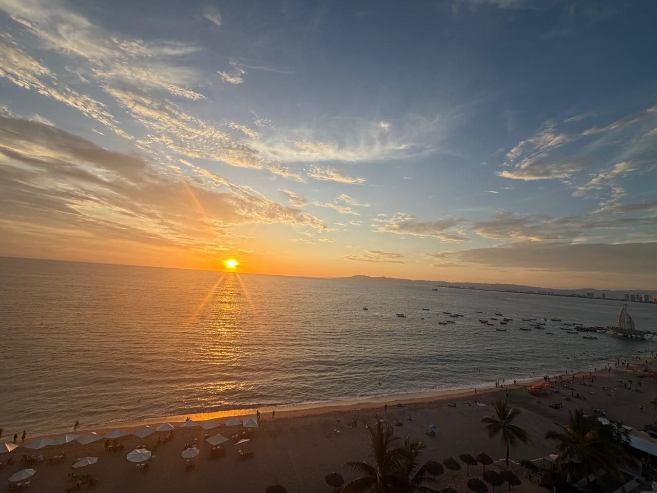 A beach scene with the sun setting over the ocean, a pier with boats, and umbrellas on the sand.
