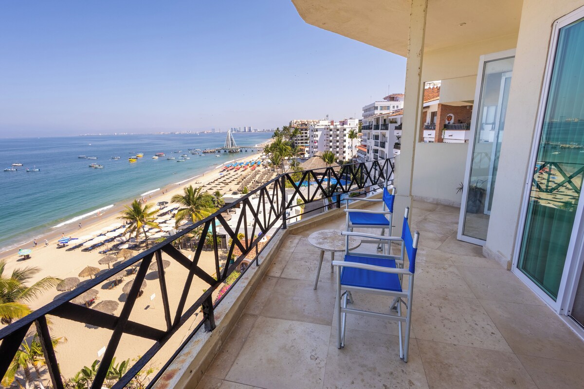A balcony with a view of a beach, a railing, and a table with chairs.