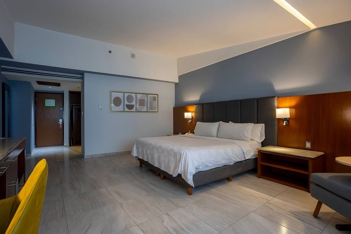 A modern hotel room with a large bed, bedside lamps, a wooden headboard, and a small wooden side table.