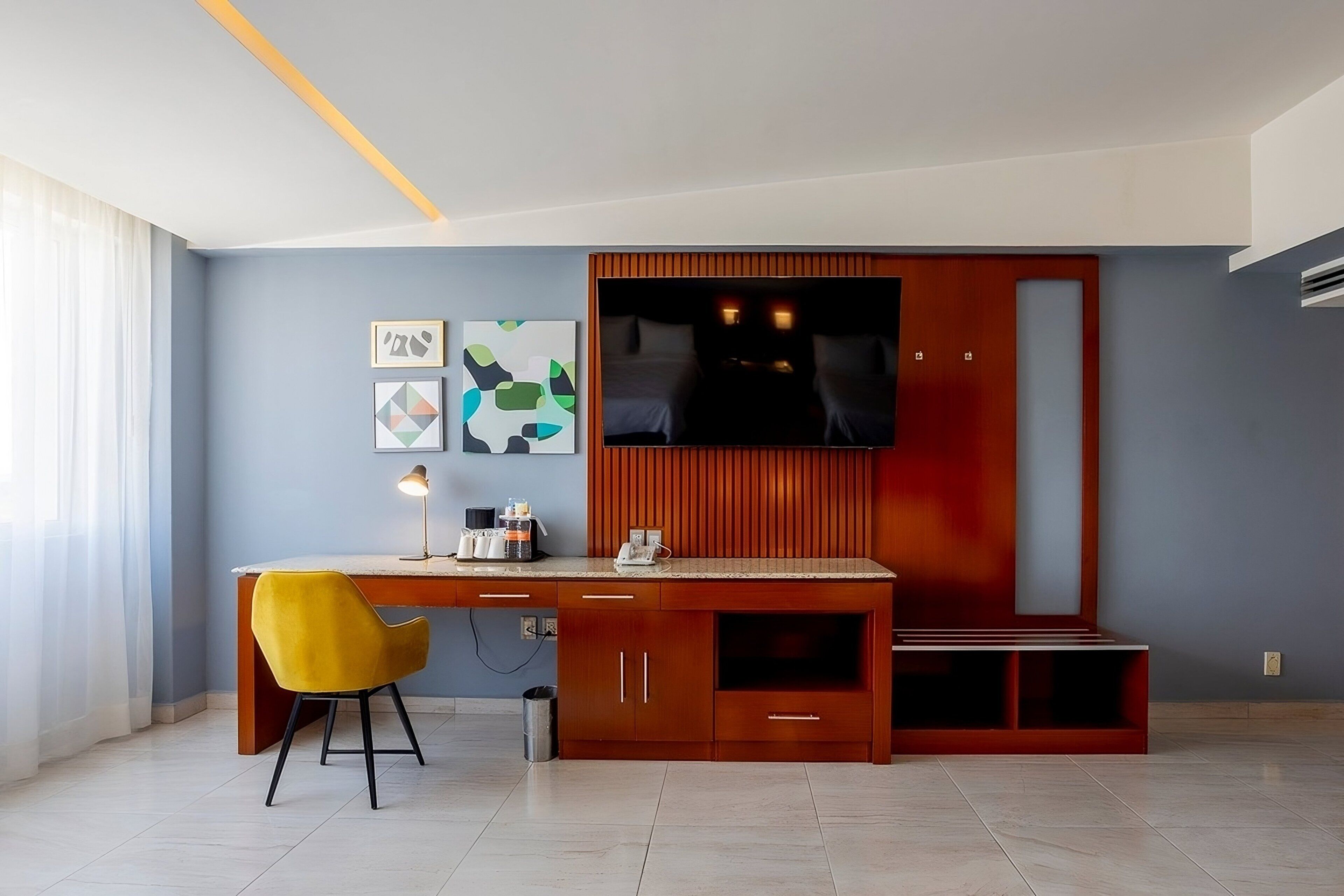 Une chambre d’hôtel moderne avec un bureau en bois, une chaise jaune, un téléviseur et des œuvres d’art accrochées au mur.