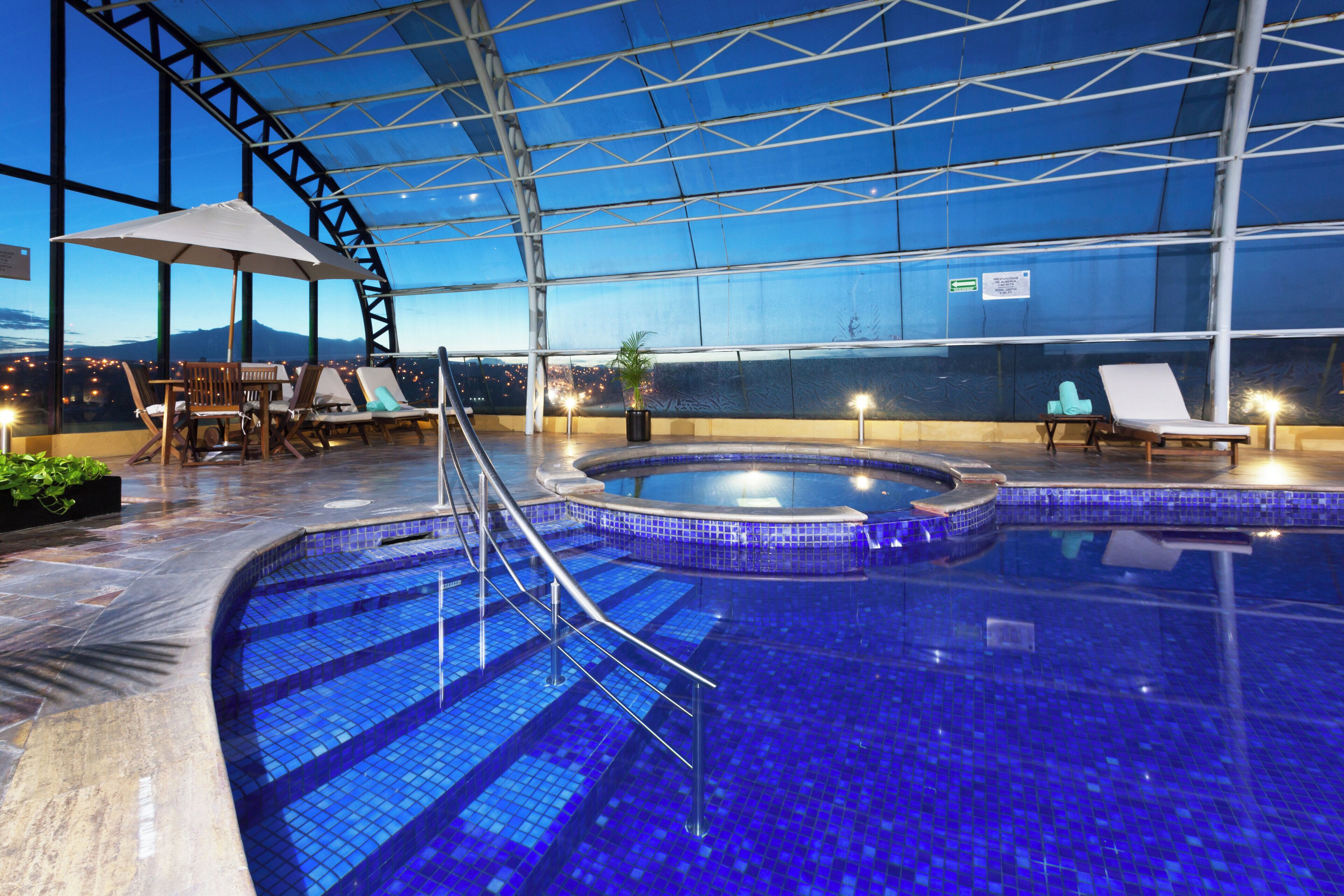 An indoor swimming pool with a glass ceiling, lounge chairs, and a city view.