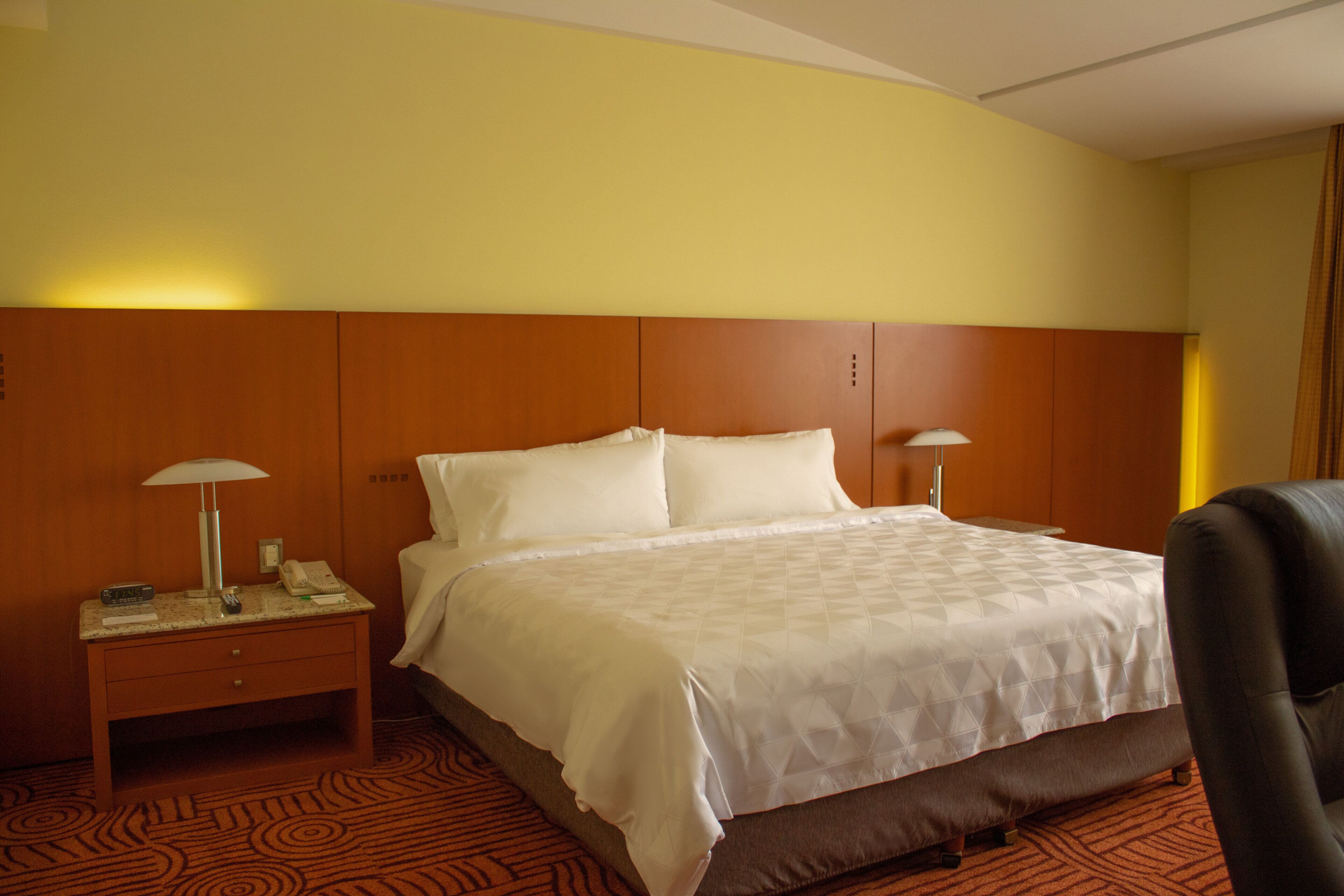 A hotel room with a bed, two bedside tables, a lamp, a chair, and a carpeted floor.