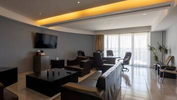 Un lobby moderno de hotel con un sofá largo, una mesa de centro, un televisor montado en la pared y un gran ventanal con cortinas.